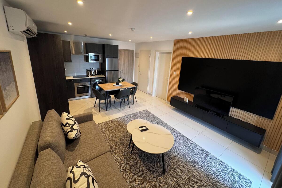 A modern living room with a sofa, coffee table, and TV. There is a kitchen area with a dining table and chairs.