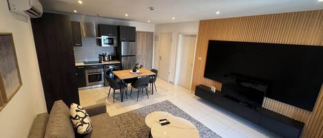 A modern living room with a sofa, coffee table, and TV. There is a kitchen area with a dining table and chairs.