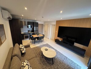 Ein modernes Wohnzimmer mit Sofa, Couchtisch und Fernseher. Es gibt eine Küchenzeile mit Esstisch und Stühlen.