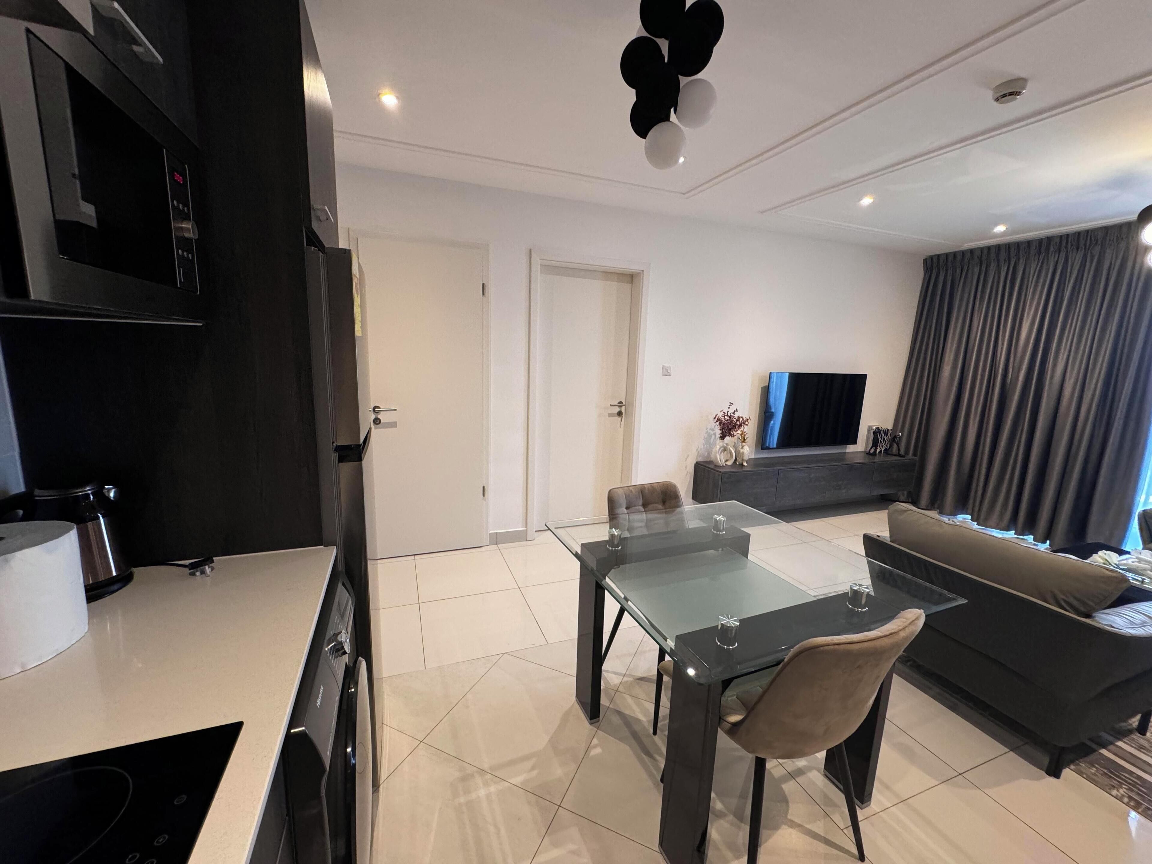 A modern living room with a glass dining table, dark sofas, and a flat-screen TV.