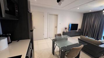 A modern living room with a glass dining table, dark sofas, and a flat-screen TV.