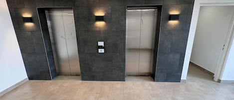 Un área de ascensores moderna con dos ascensores, luminarias fijadas a la pared y un letrero de salida de emergencia verde.