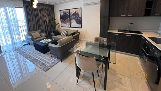 A modern living room with a sofa, coffee table, and a glass dining table. There is a kitchen area with dark cabinets and a sink. - Loxwood Abode (Accra)