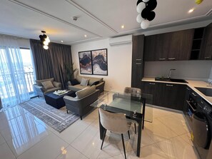 Een moderne woonkamer met een bank, salontafel en een glazen eettafel. Er is een keuken met donkere kastjes en een spoelbak.