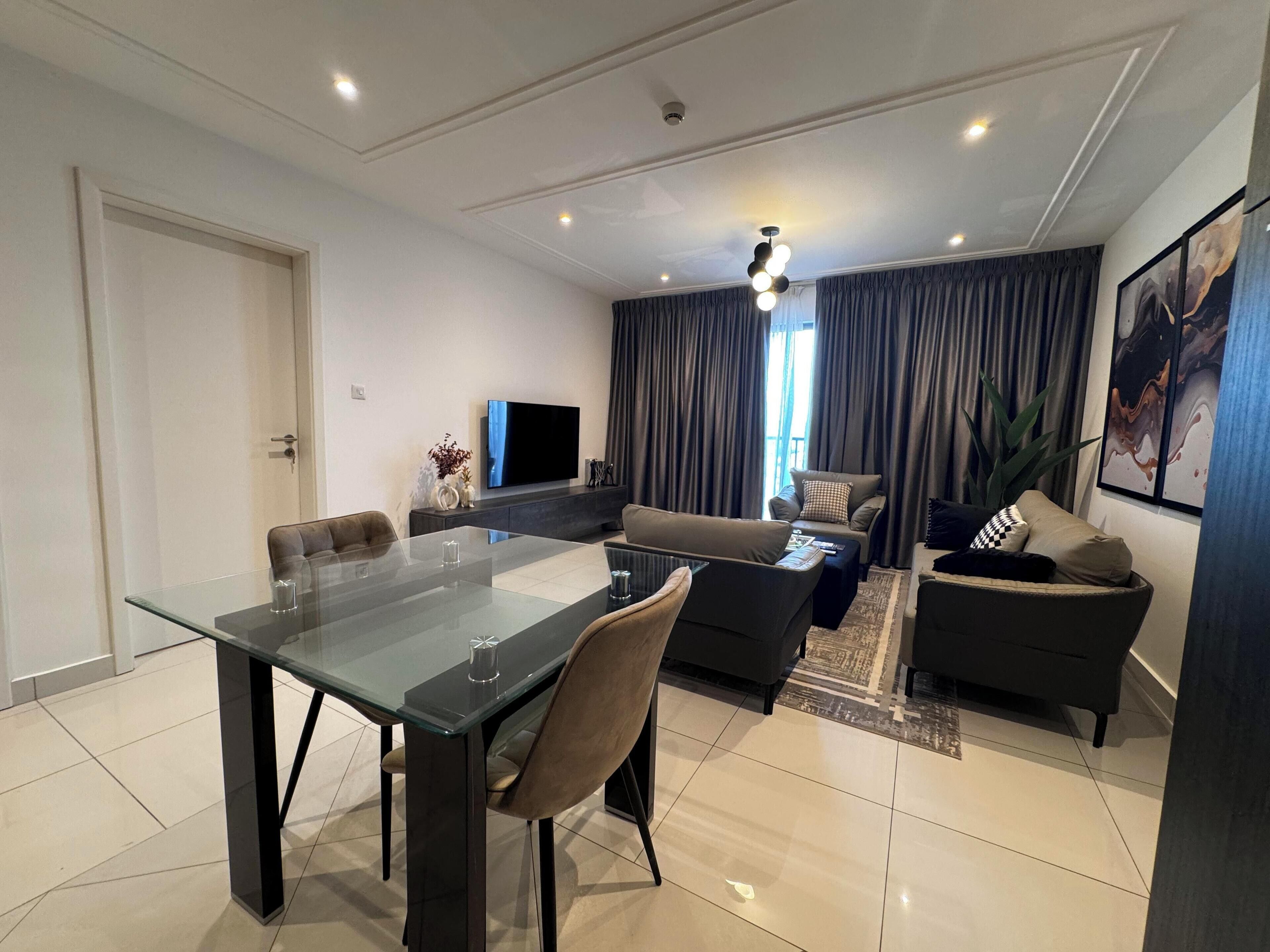 Una sala de estar moderna con una mesa de comedor de cristal, sofás, un televisor de pantalla plana y un ventanal con cortinas.