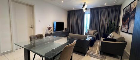 Un salon moderne avec une table à manger en verre, des canapés, un téléviseur à écran plat et une grande fenêtre avec des rideaux.