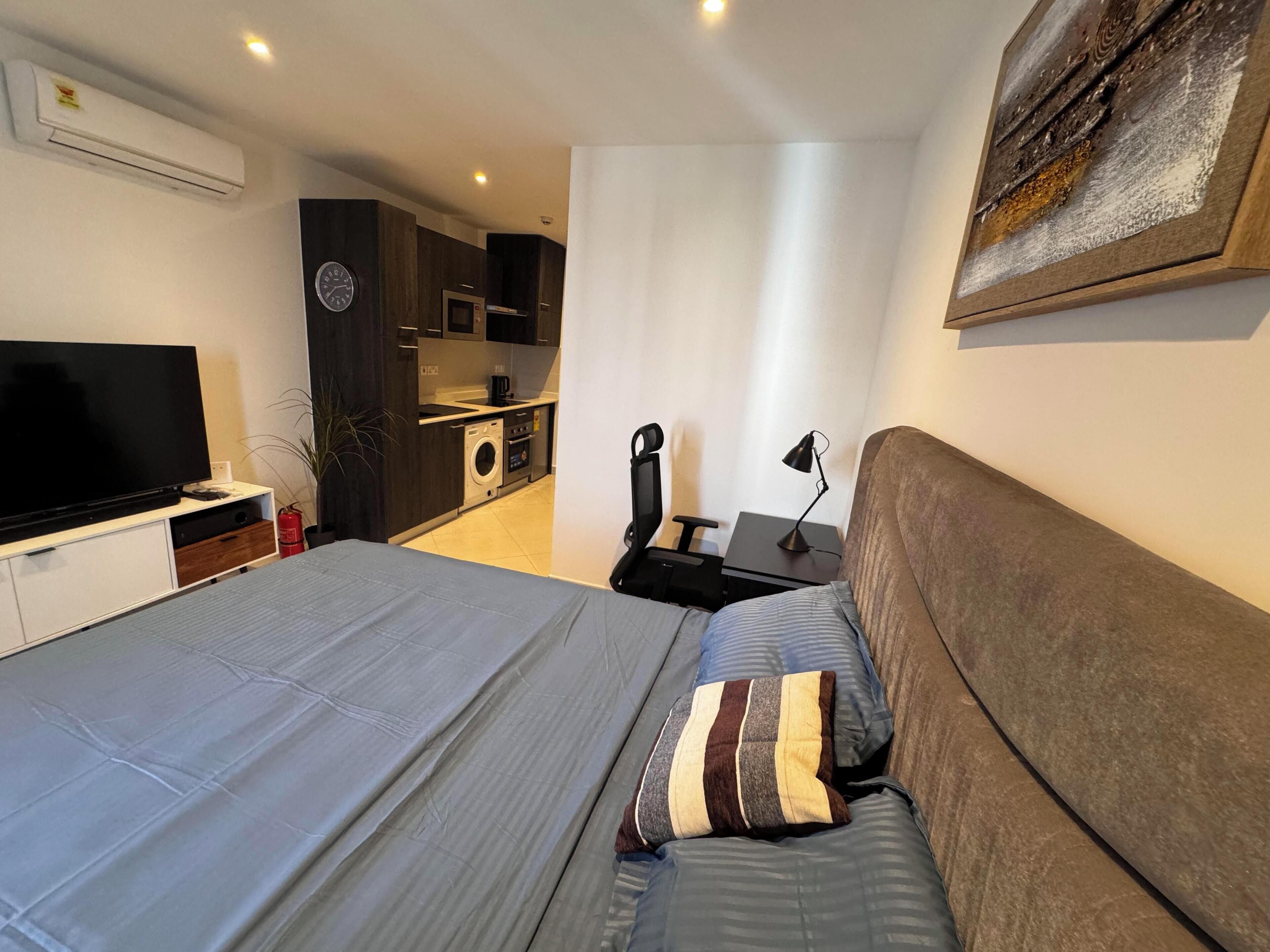 A bedroom with a bed, a TV, a desk, and a kitchenette.