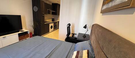 A bedroom with a bed, a TV, a desk, and a kitchenette.