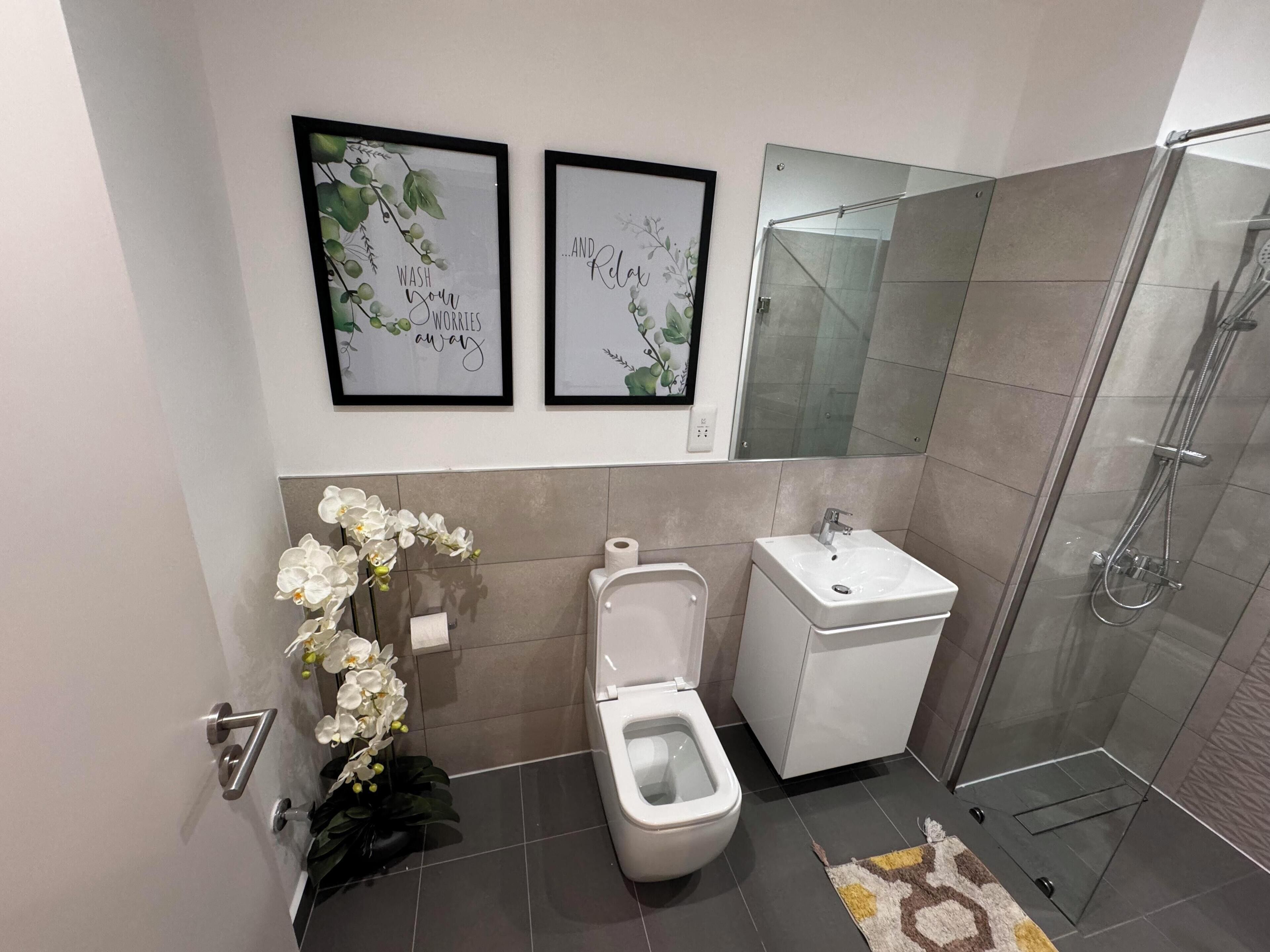 Un baño moderno con inodoro, lavamanos y cabina de ducha de vidrio. Dos obras de arte enmarcadas decoran la pared sobre el lavamanos.