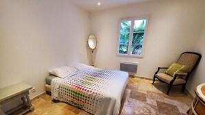 A bedroom with a bed, a chair, a small table, a window with a view of greenery, and a radiator. - Villa Azur 6 bedrooms and heated pool 20 persons (Le Thoronet)