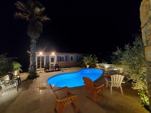 Una piscina con sillas y una mesa, rodeada de plantas y un edificio con luces.