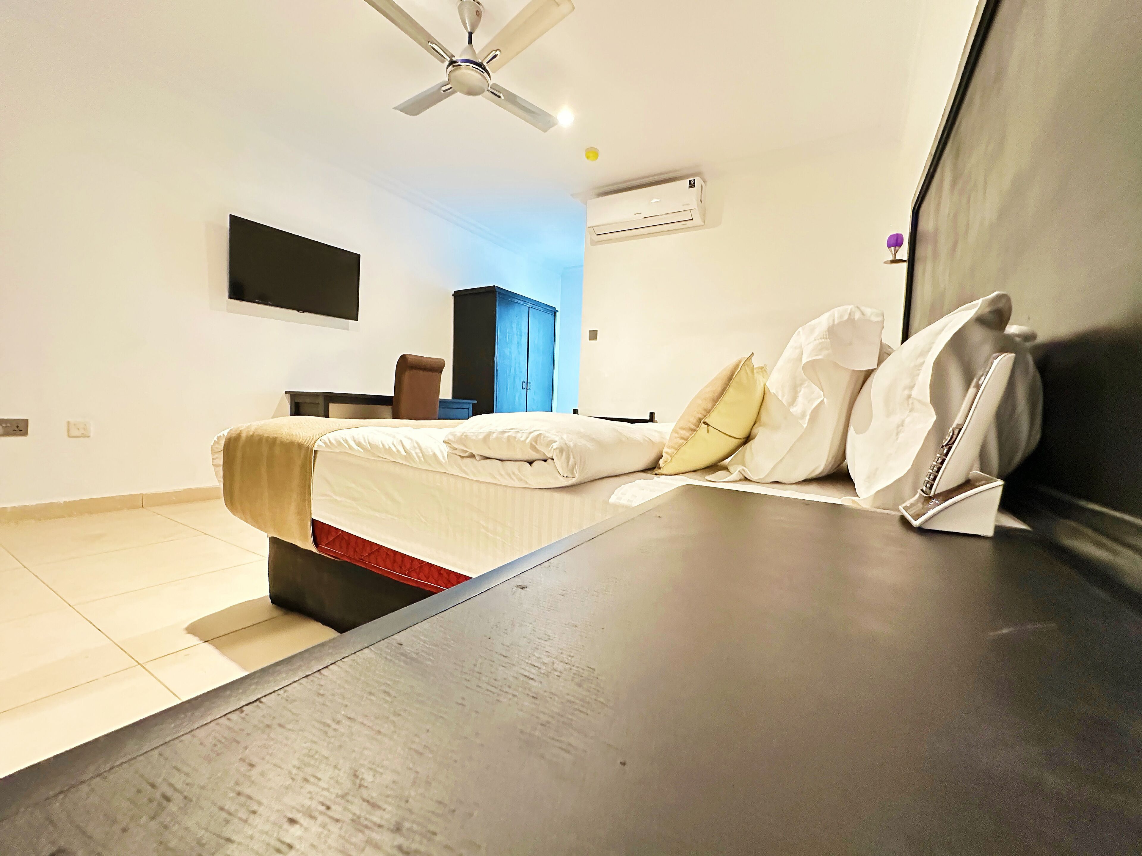 Un dormitorio moderno con una cama, ventilador de techo, unidad de aire acondicionado, televisor montado en la pared y un armario oscuro.