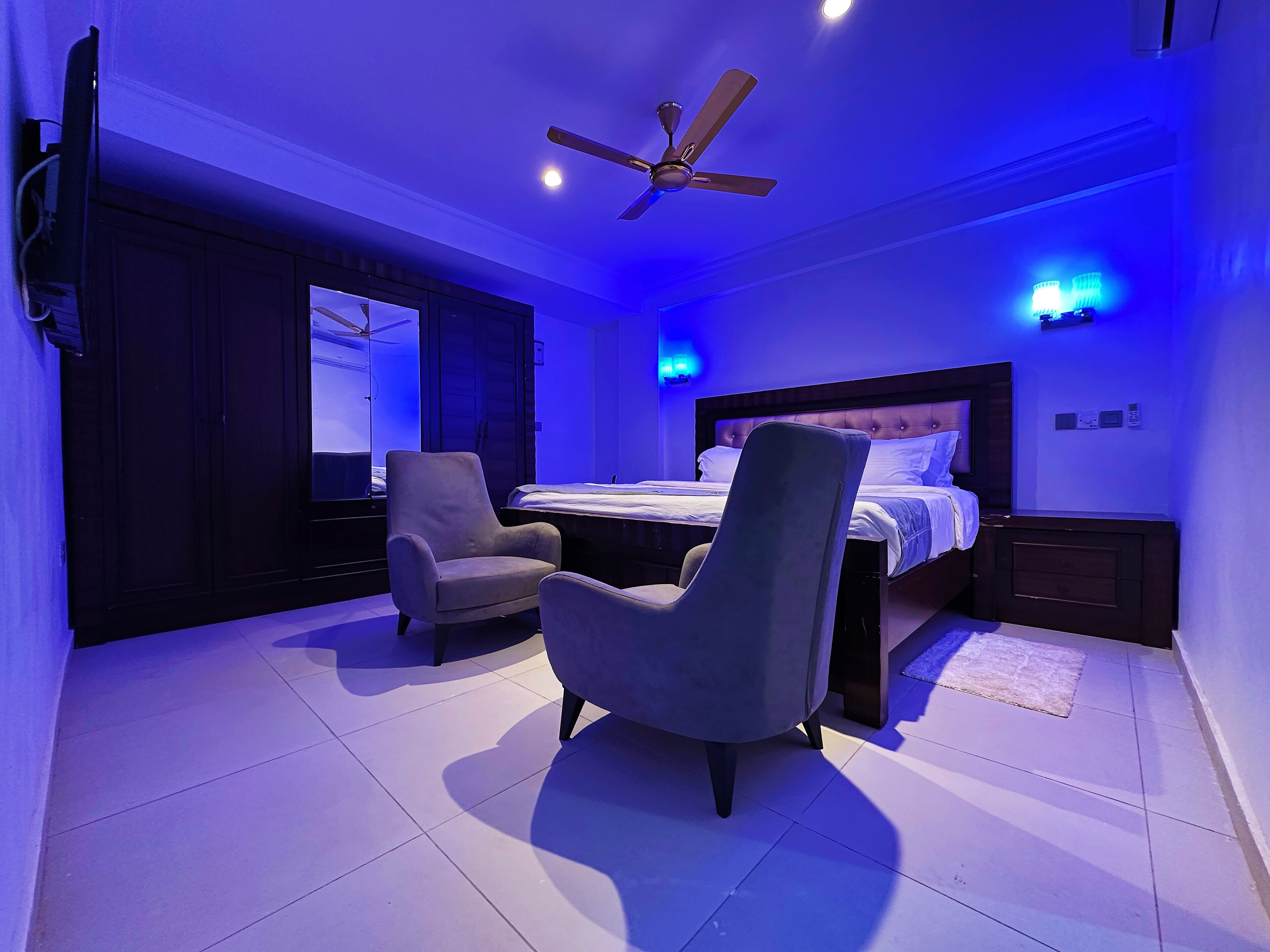 Un dormitorio con cama, dos sillones, ventilador de techo e iluminación ambiental azul.