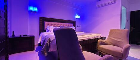 Een slaapkamer met een bed, een plafondventilator, een nachtkastje, een stoel en een wandairconditioner.