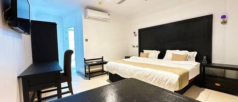 A hotel room with a bed, bedside table, desk, chair, ceiling fan, and an air conditioning unit.