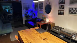 Ein gemütliches Wohnzimmer mit einem Holzespeisetisch, einer schwarzen Ledersofa und einem kleinen Couchtisch. Es gibt Blumentöpfe, ein gerahmtes Bild und eine Lampe auf dem Tisch.