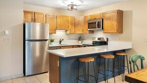 A kitchen with wooden cabinets, a stainless steel refrigerator, a microwave, and a bar area with stools. - 6 Mi to Skiing: Condo w/ Balcony in Fraser! (Fraser)