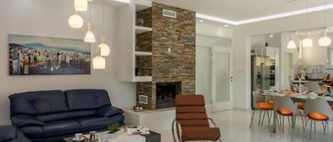 A modern living room with a sofa, armchairs, and a coffee table. There is a fireplace and a dining area visible in the background.