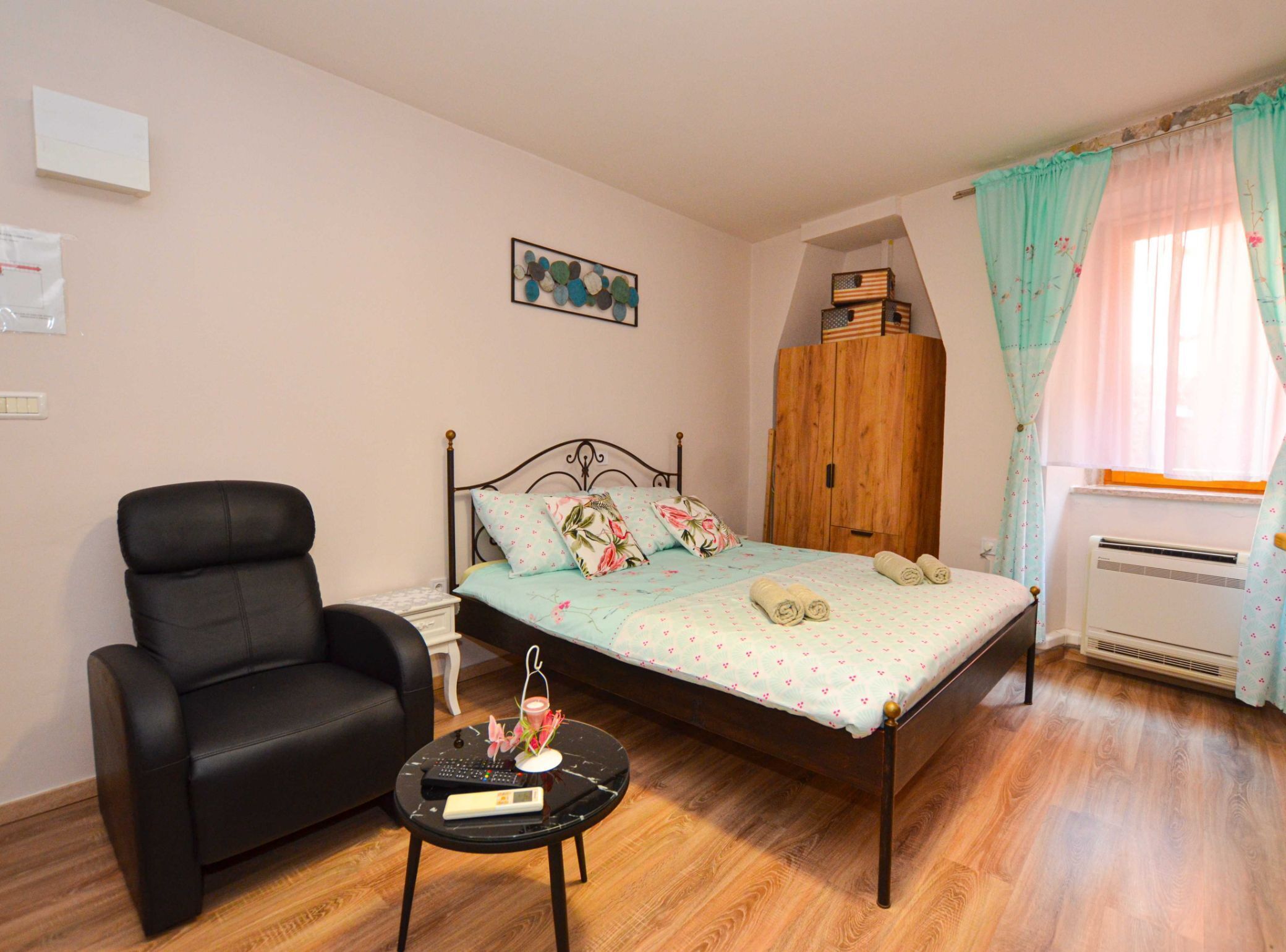 A bedroom with a bed, a chair, a small table, a wardrobe, and a window with curtains.
