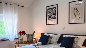Une chambre d’hôtel avec un lit, des oreillers, une table de chevet ornée d’un vase de fleurs, un seau et une œuvre d’art encadrée accrochée au mur.