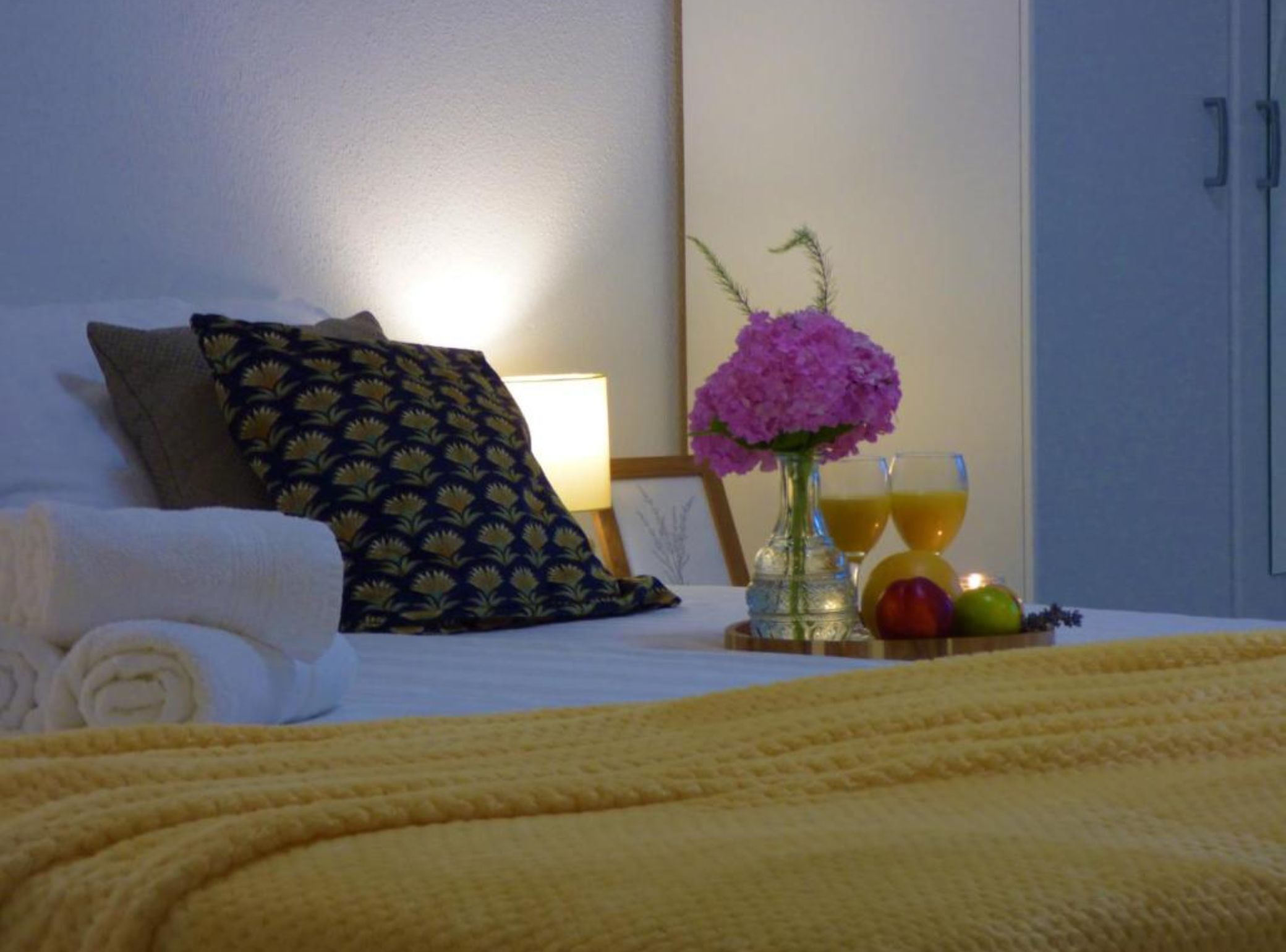 A neatly made bed with a yellow blanket, a dark patterned pillow, a vase with pink flowers, and a fruit bowl with oranges and apples.