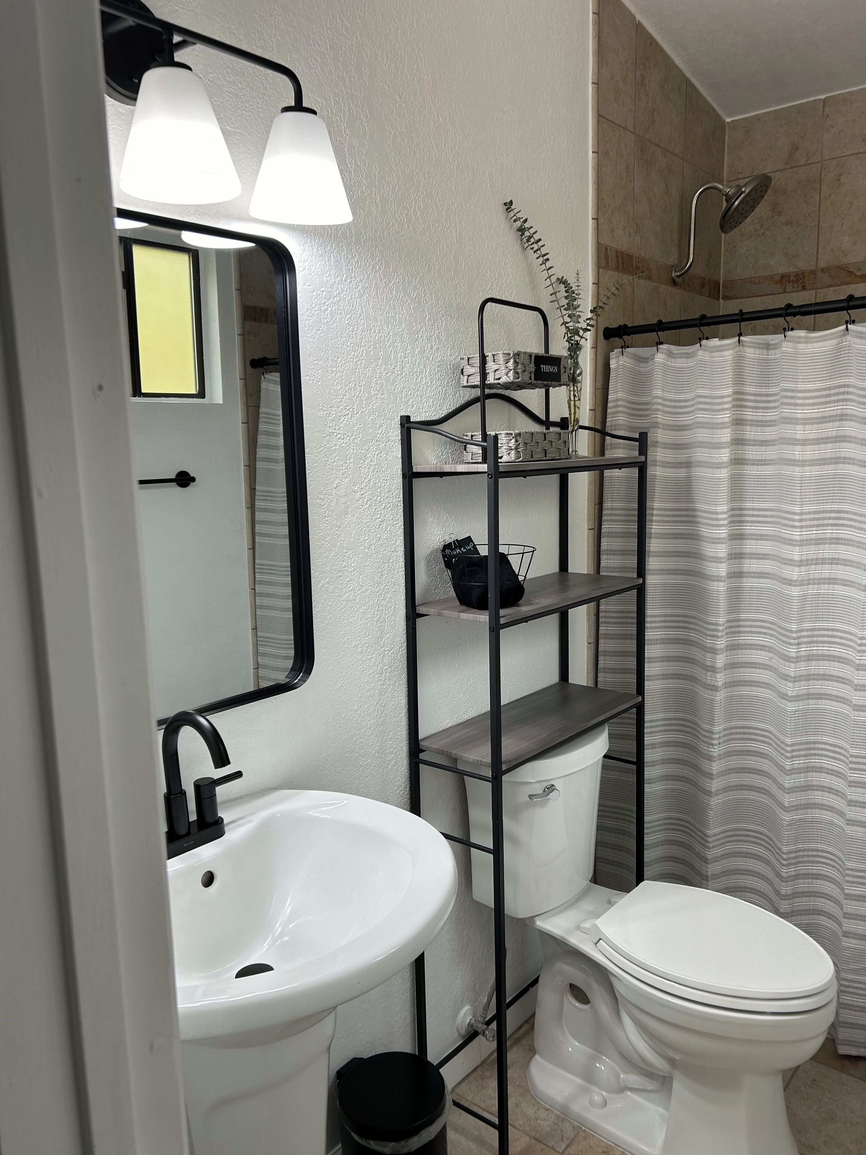 Et badeværelse med en hvid vask, hane i sort farve, toilet, brusebad med kurvet brusehoved og et stribet brusebadeforhæng. Der er et spejl med sort ramme og en hylde med en plante og en sort taske.