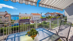 A balcony with a table and chairs, a bowl of green fruits, and a view of the sea and houses. - Modern Crikvenica Apartments With Sea View (Crikvenica)