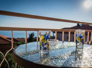 Een glazen tafel met twee glaasjes met blauwe en gele bloemmotieven, voor een balkonhek met een helder uitzicht op de lucht en verre gebouwen.
