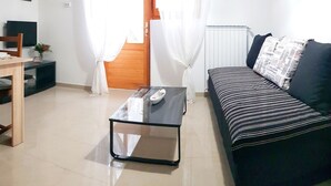 A living room with a black sofa, a glass coffee table, a wooden dining table, and a TV. There are two framed pictures on the wall, a clock, and a window with white curtains. - Seafront Chic Crikvenica Apartments (Crikvenica)