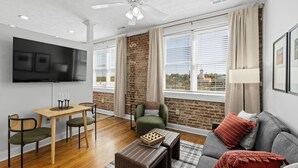 Una sala de estar moderna con un televisor de pantalla plana, un comedor con mesa y sillas, un sofá con cojines y ventanas amplias con cortinas.