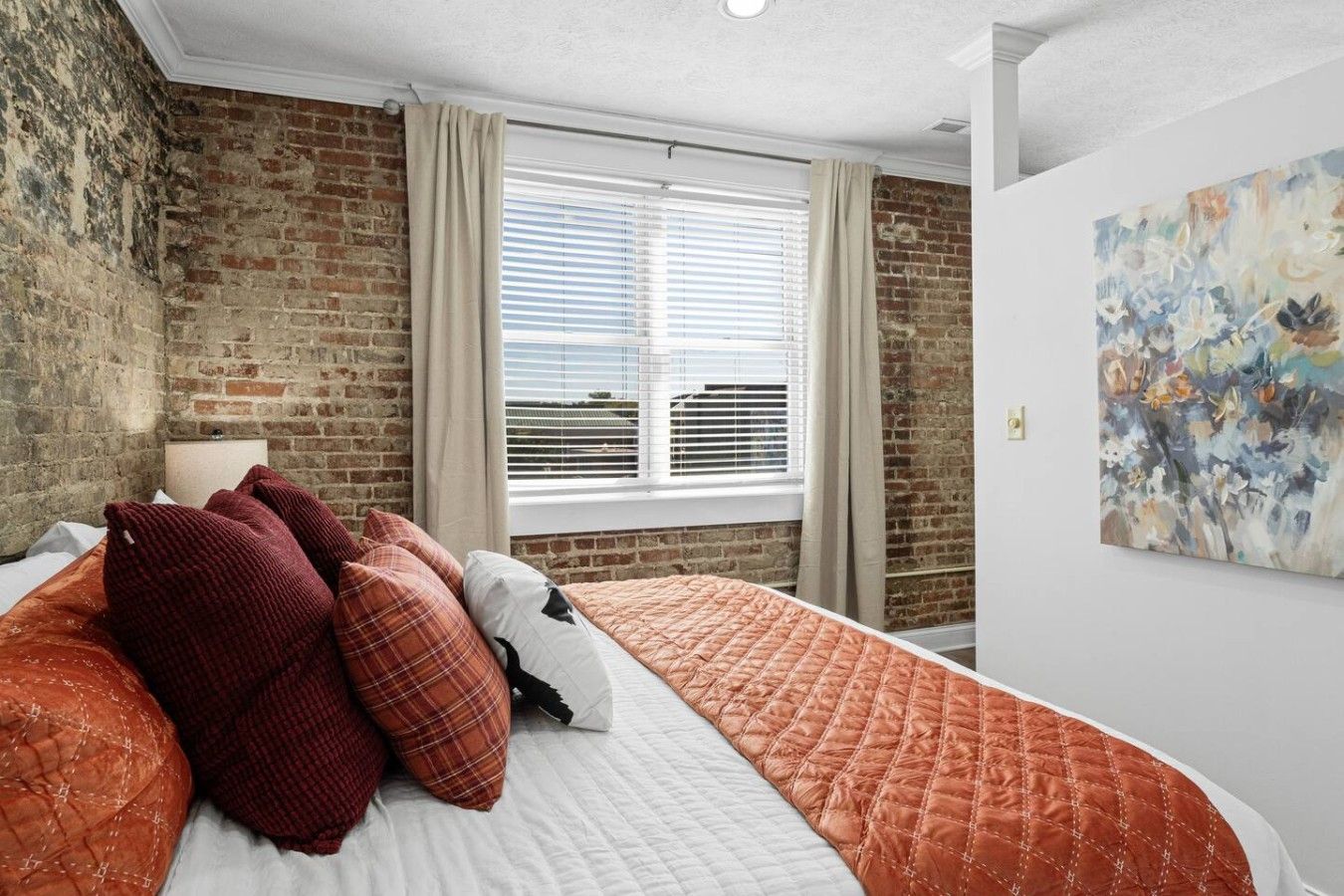 A bedroom with a brick wall, a bed with a quilted cover, a window with blinds, and a painting on the wall.