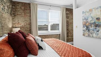 A bedroom with a brick wall, a bed with a quilted cover, a window with blinds, and a painting on the wall.