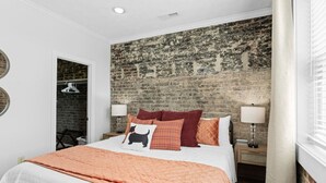 Un dormitorio con pared de ladrillo, una cama con varios cojines, dos mesitas de noche con lámparas y un armario.