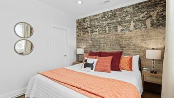 A bedroom with a brick wall, a white door, two round mirrors, a bed with multiple pillows, and bedside lamps.