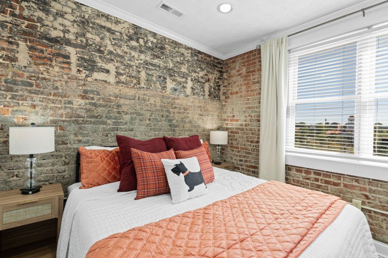 A bedroom with a brick wall, a bed with multiple pillows, two bedside lamps, and a window with blinds.
