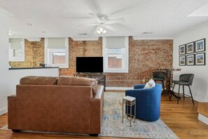 A living room with a brown sofa, a blue armchair, a TV, and a brick wall. - Blue Bella a Downtown Maryville Loft (Maryville)