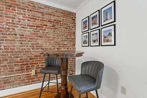 Una habitación con pared de ladrillo, una mesa redonda de madera con dos sillas y seis cuadros en la pared.