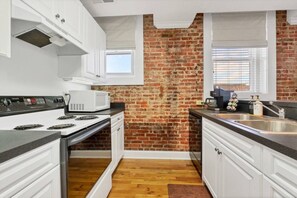 Una cocina con armarios blancos, pared de ladrillo, microondas, estufa y lavaplatos.