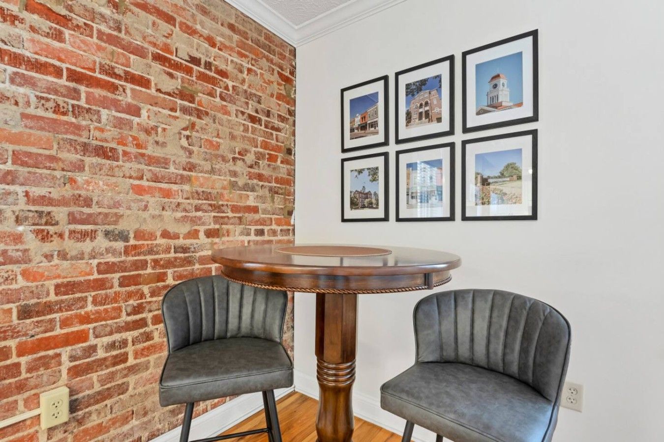A room with a brick wall, a round wooden table, two grey upholstered chairs, and six framed pictures on the wall.