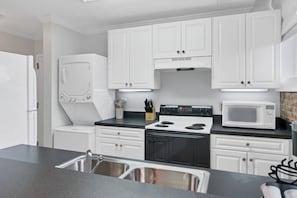 Una cocina con armarios blancos, una estufa negra, un microondas y un lavamanos de acero inoxidable.