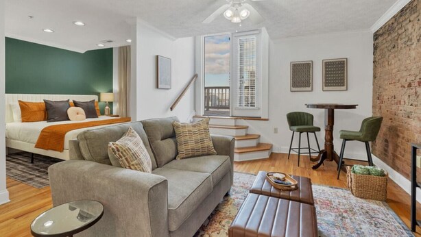 Ein modernes Wohnzimmer mit Sofa, Couchtisch und Essbereich mit Stühlen. Eine Treppe führt auf eine obere Ebene, ein großes Fenster mit Jalousien und ein Deckenventilator sind ebenfalls vorhanden.