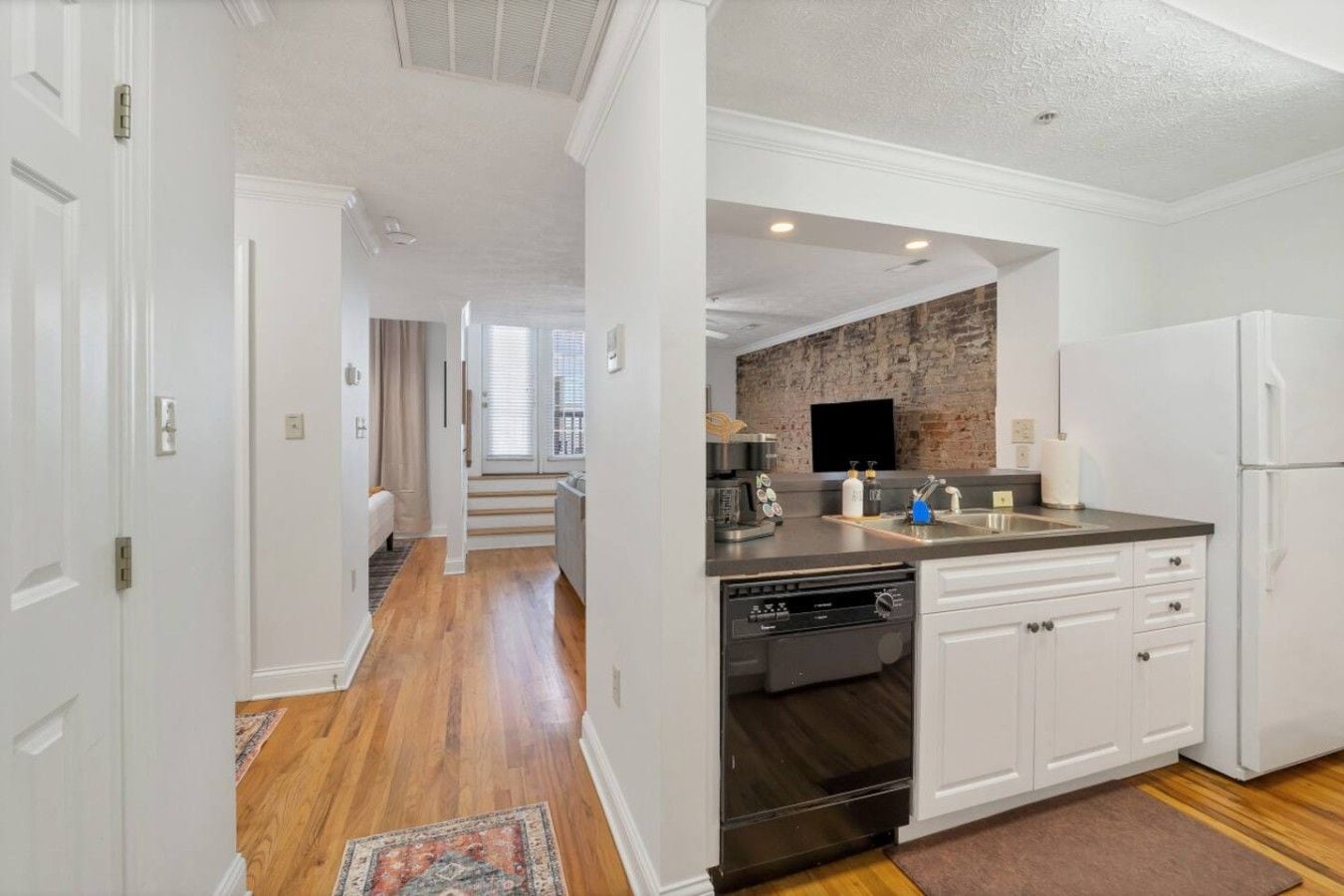 A modern kitchen with white cabinetry, a black dishwasher, and a brick accent wall.