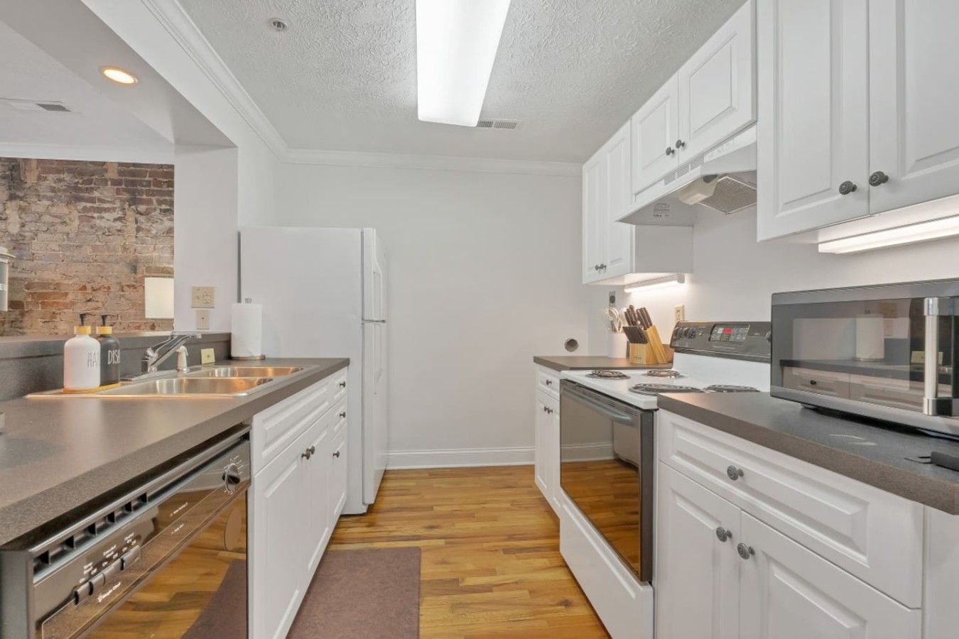 A modern kitchen with white cabinetry, a built-in oven, a microwave, and a brick accent wall.
