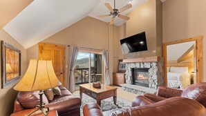 A spacious living room with leather sofas, a fireplace, a ceiling fan, and a TV mounted on the wall.