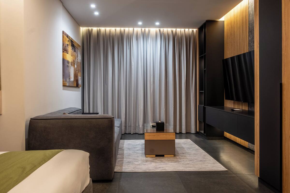 A modern hotel room with a large bed, a small sofa, a wooden side table, a flat-screen TV, and a large window with curtains.