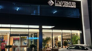 Het exterieur van L'Avenue Hotel Apartments, met een modern ontwerp, grote glasramen en een geparkeerde auto ervoor.
