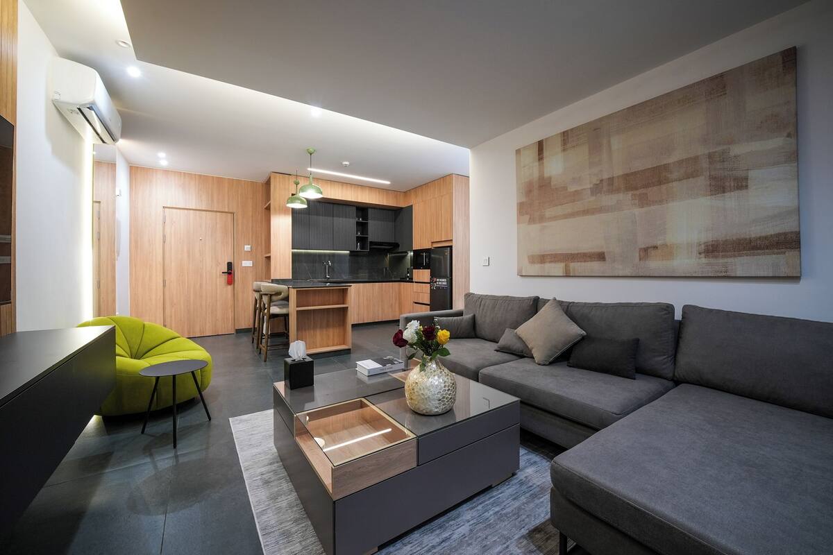 A modern living room with a sofa, a coffee table, and a bright green chair. There is a kitchen area with wooden cabinets and a black countertop.