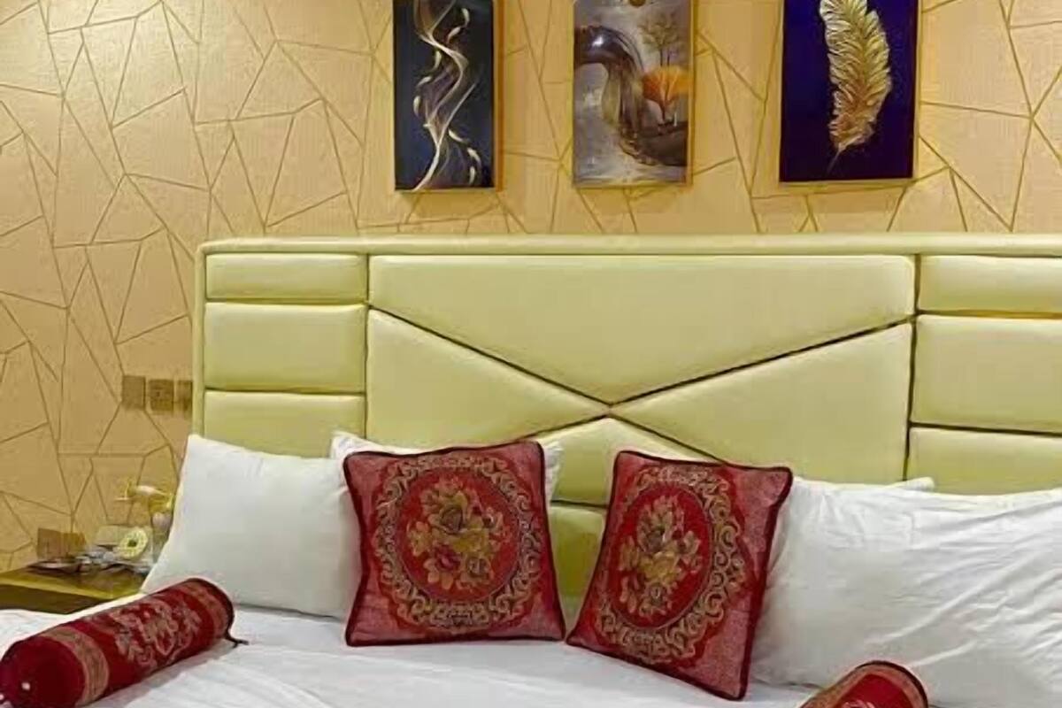 A hotel room with a geometric-patterned wall, a light green upholstered headboard, and three artworks above the bed.