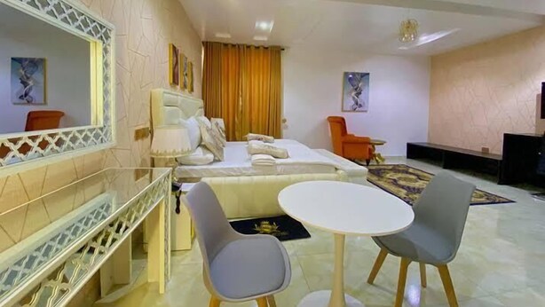 A hotel room with a bed, a desk, two chairs, a small table, and a TV. - Hogis Luxury Suites and Hotel (Calabar)