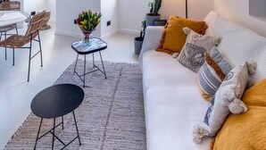 Una sala de estar moderna con un sofá blanco, una mesa de comedor y una mesa de centro. Hay un televisor montado en la pared y una lámpara en una mesita auxiliar.